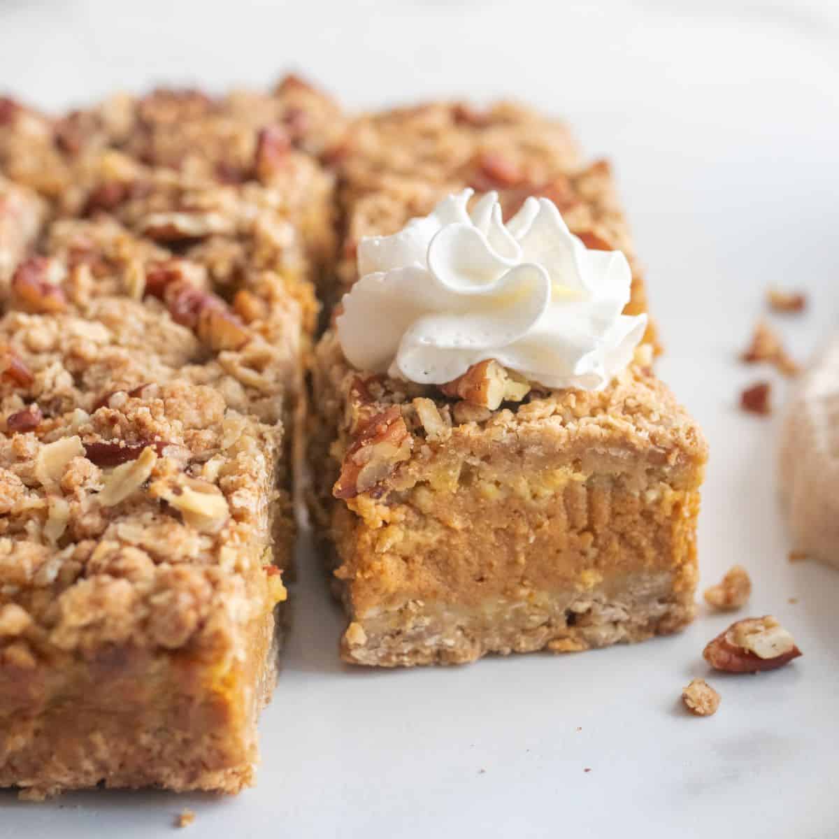 Cut pumpkin pecan bars on a counter top, focused on one topped with maple whipped cream.