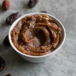 a bowl of date paste topped with flaky salt on a countertop with a few medjool dates beside the bowl.