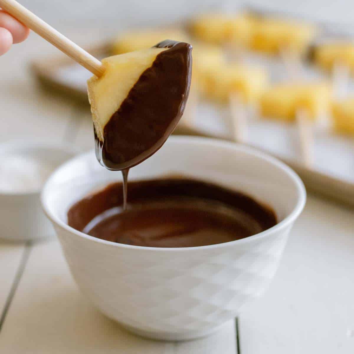 Pineapple slice on a candy apple stick dipped into a bowl of melted chocolate.