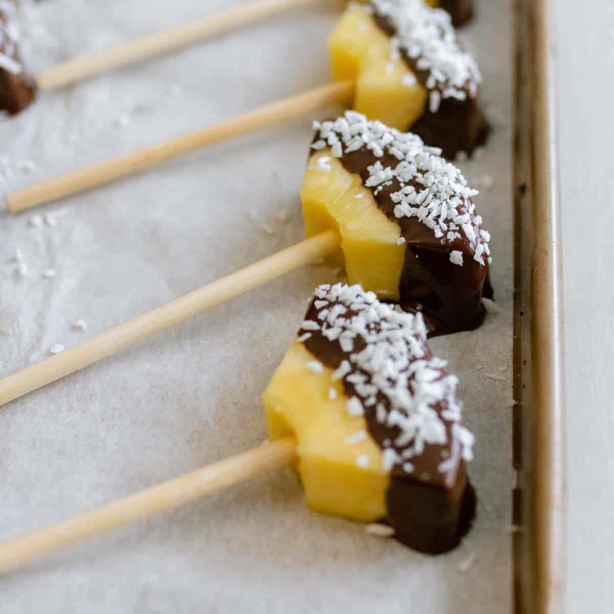 Chocolate covered pineapple dipped in shredded coconut on candy apples sticks laid on a baking sheet.