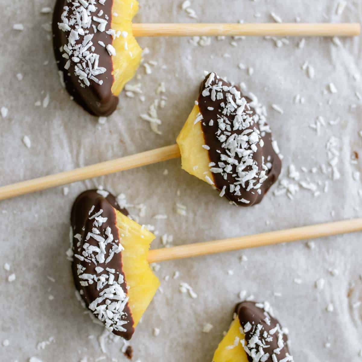 Chocolate-covered pineapples on a baking sheet with shredded coconut laying around them.