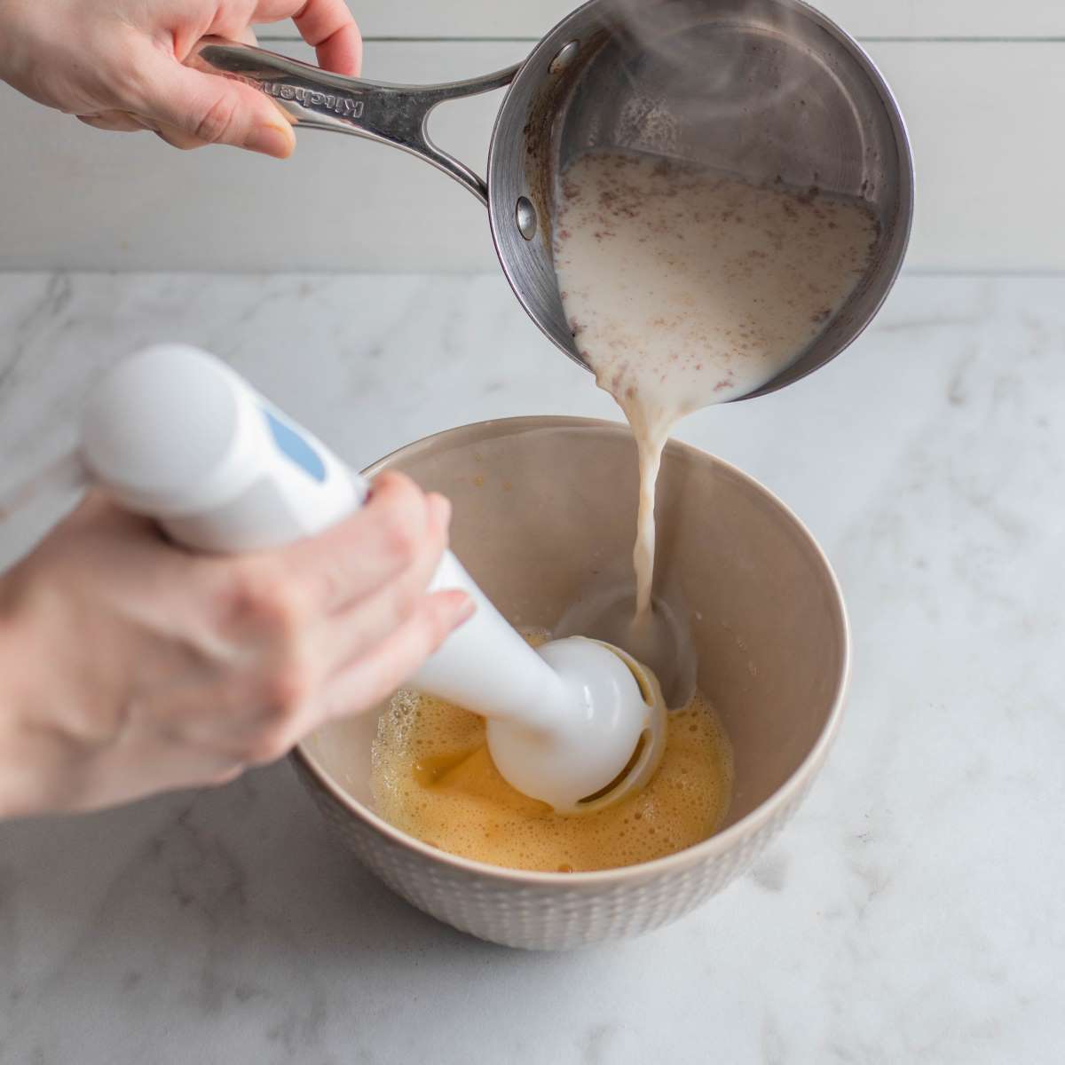 Wet ingredients are poured from a saucepan into the mixing bowl containing the whisked eggs.