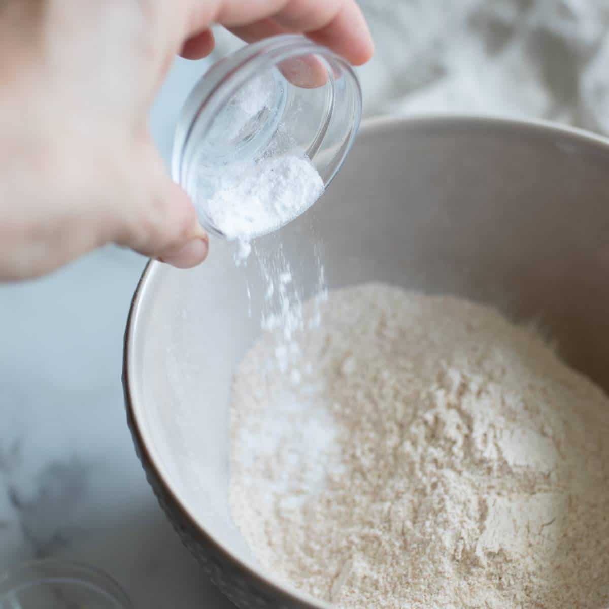 Pouring sea salt into a mixing bowl of spelt flour and baking soda.
