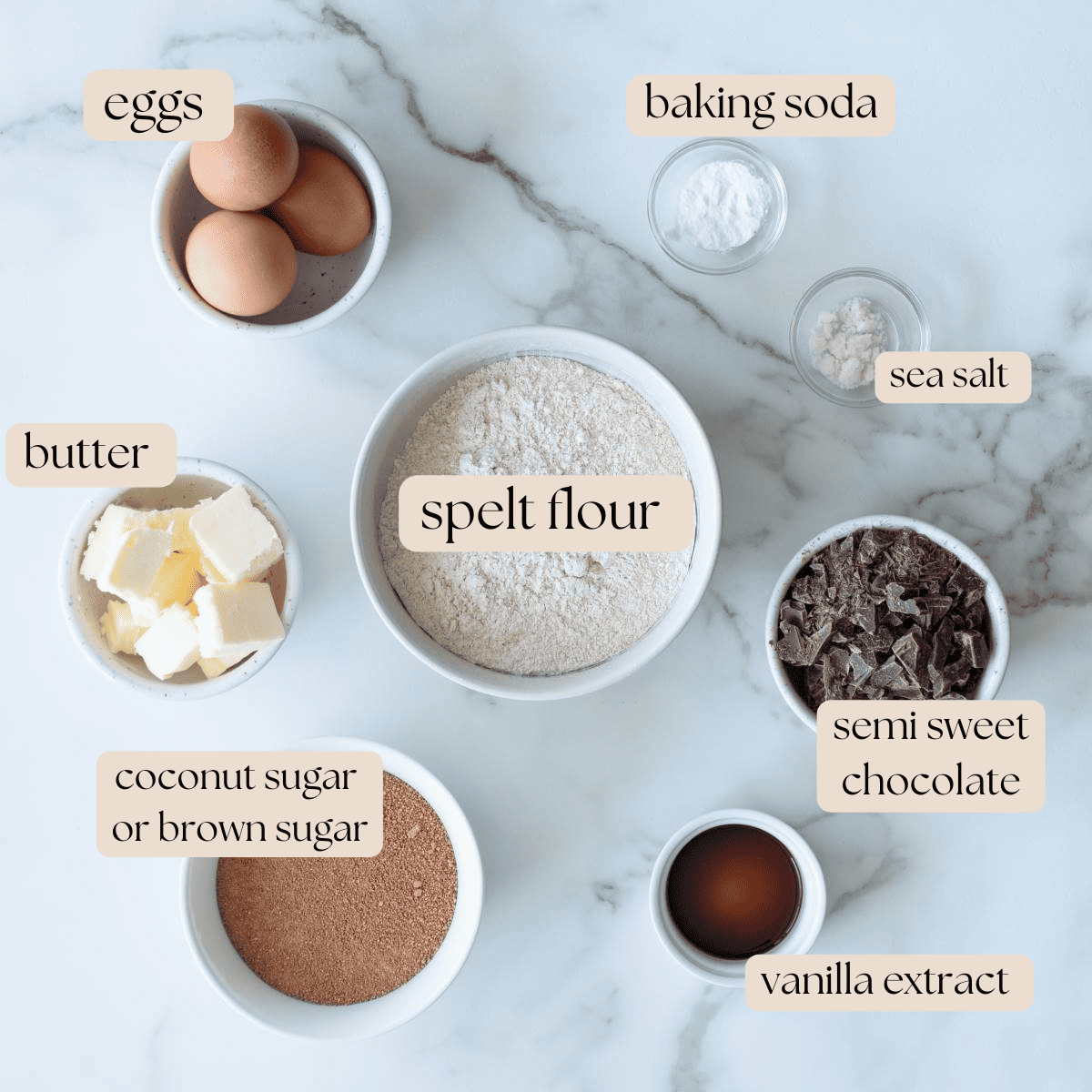 Ingredients to make spelt cookies in mixing bowls and pinch bowls on a marble countertop.