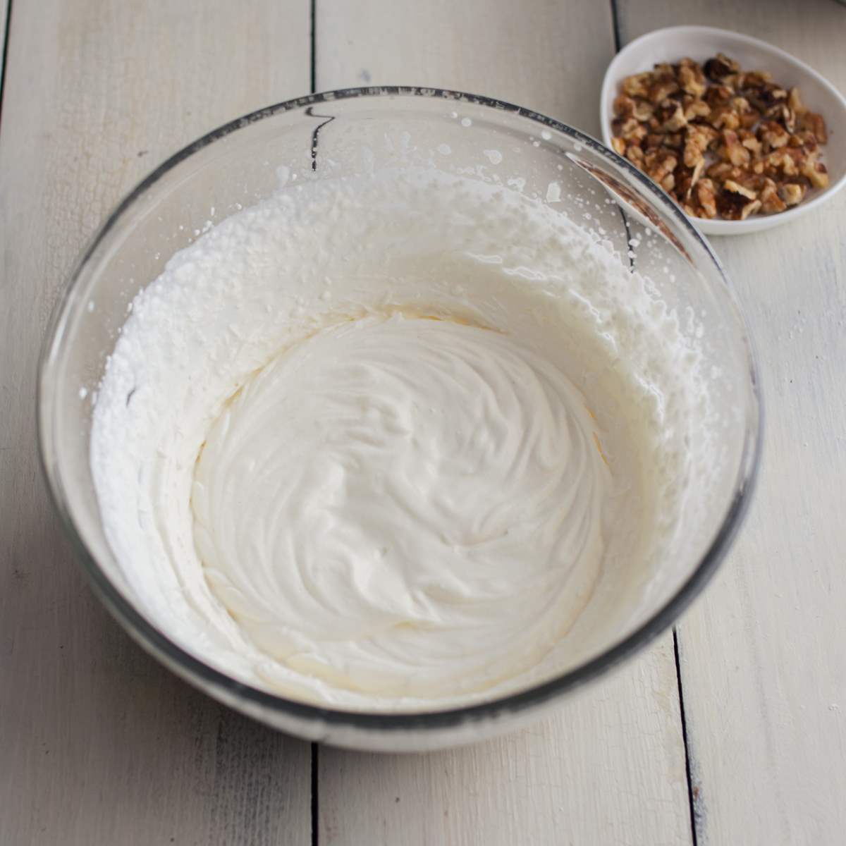 Whipped cream in a clear mixing bowl on a wooden board.