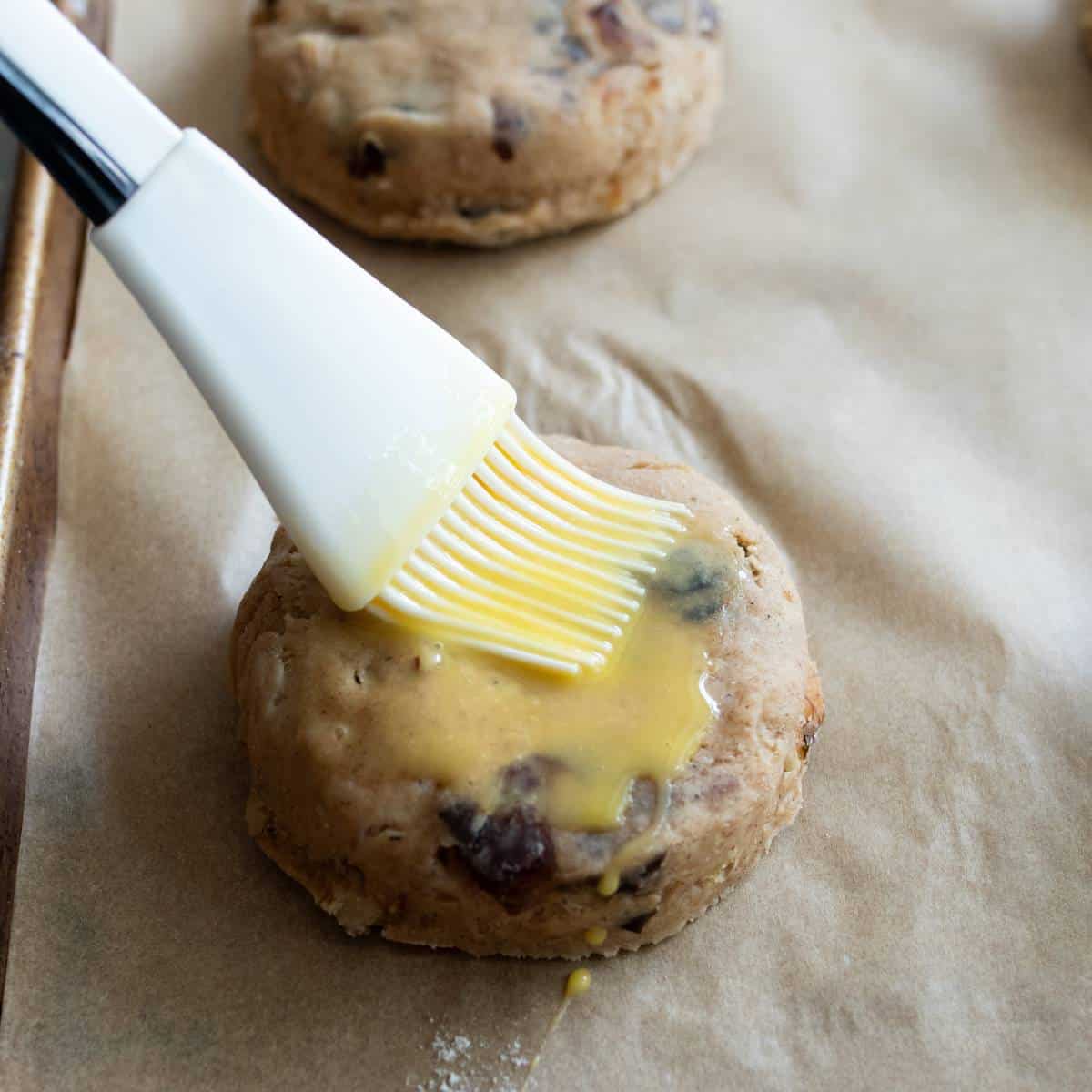 A brush adds egg wash to a scone round on a baking sheet of dough rounds before going into the oven.