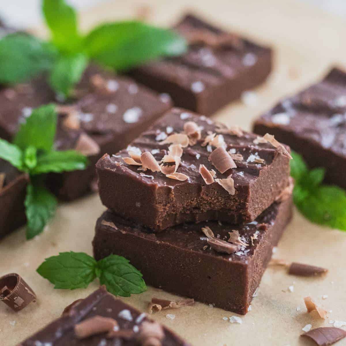 Two healthy fudge bars with chocolate and salt garnish, stacked on top of each other on a baking sheet full of bars and peppermint leaves.