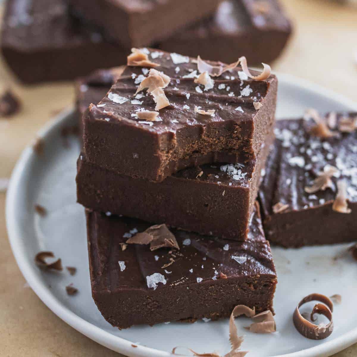 3 healthy fudge bars stacked on top of each other on a dessert dish with chocolate shavings and flaky salt sprinkled around.