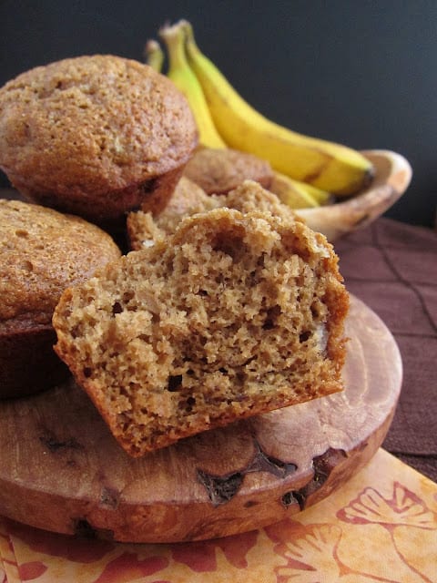 Barley banana muffins on a wooden plate with a bite taken out of the first one and bananas in a bowl behind.