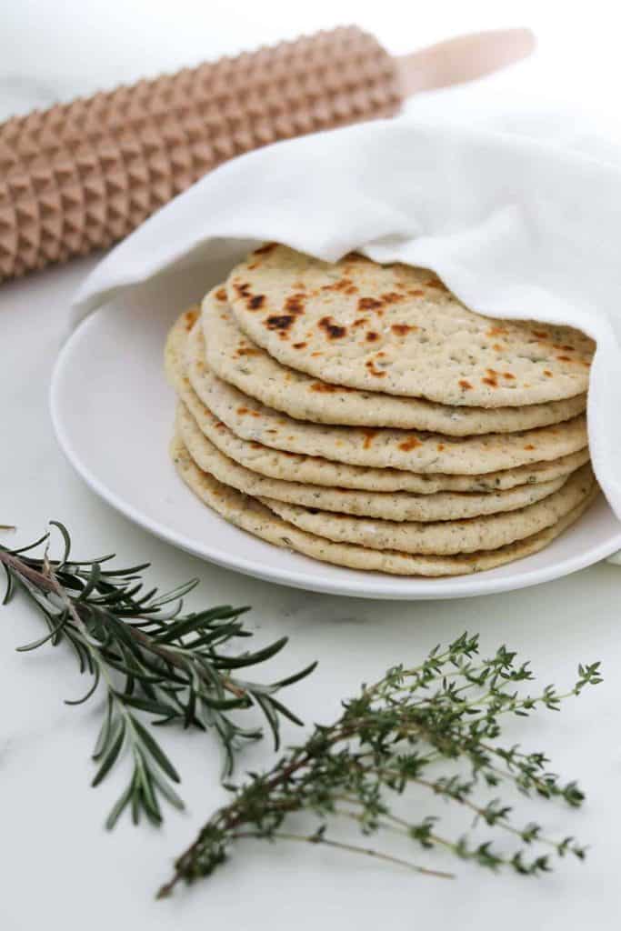 Seven Swedish flatbreads on a white plate with a napkin on top and garden herbs nearby.