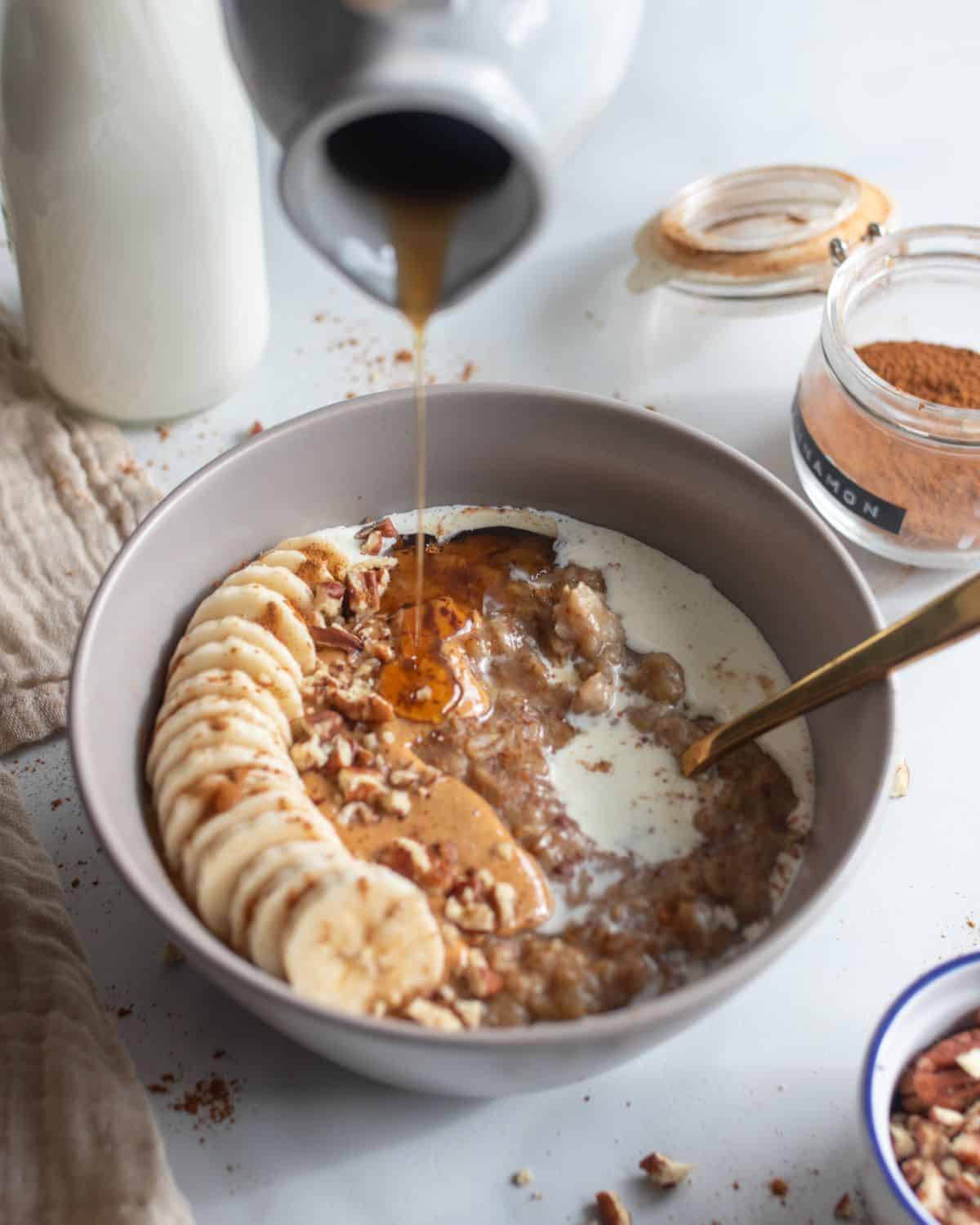 Bowl of date oatmeal with banana, pecan and cream topping, with maple syrup being poured over it.