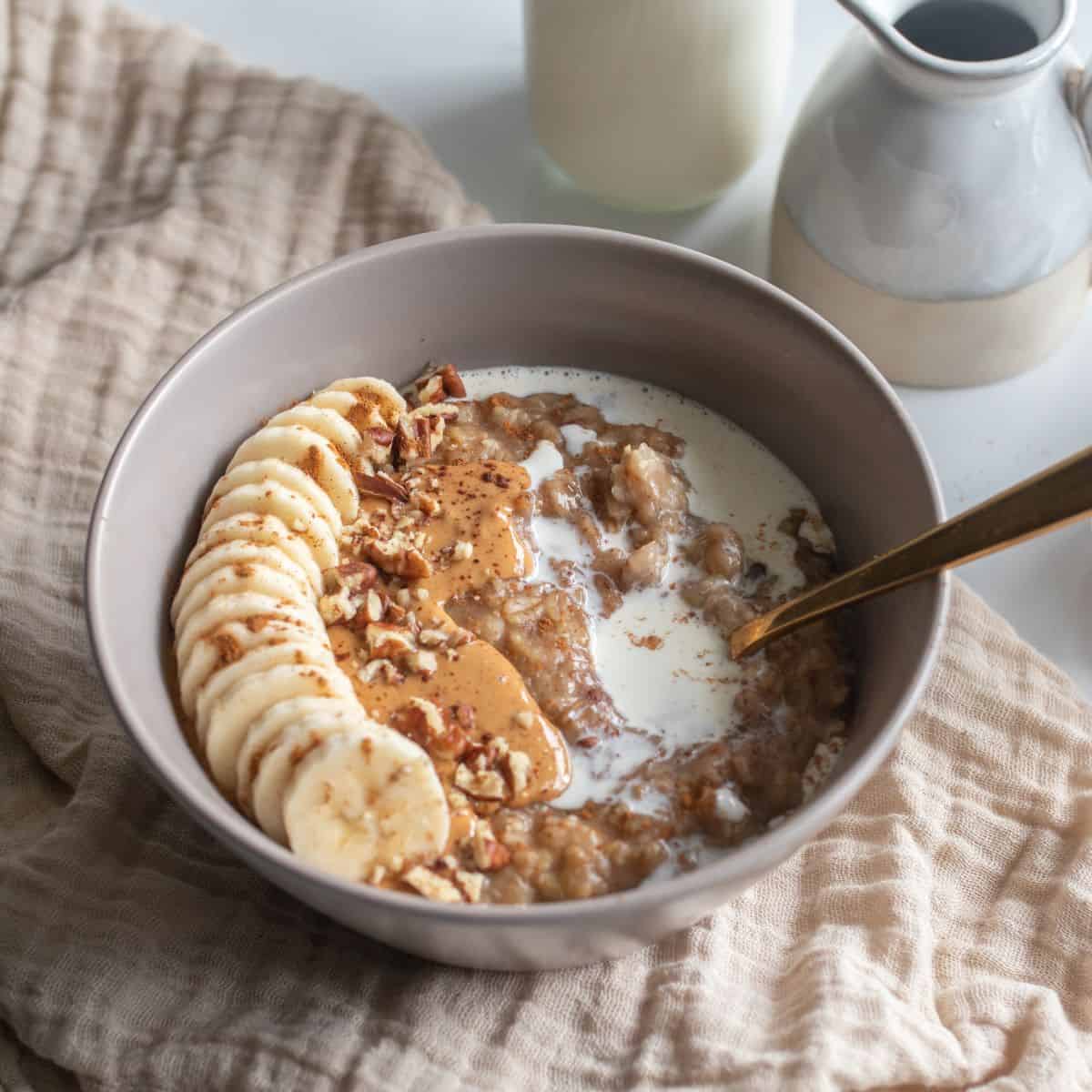 Bowl of date oatmeal with banana, pecan and cream topping on a staged table top.