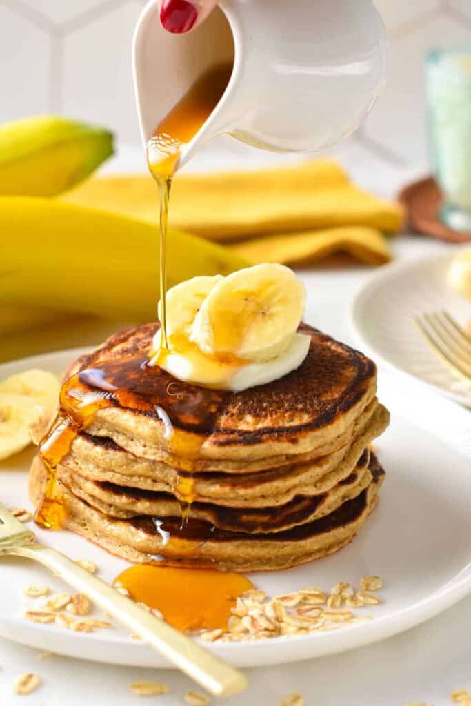 Four pancakes stacked on top of each other on a place with oats and banana slices around and maple syrup pouring on top.