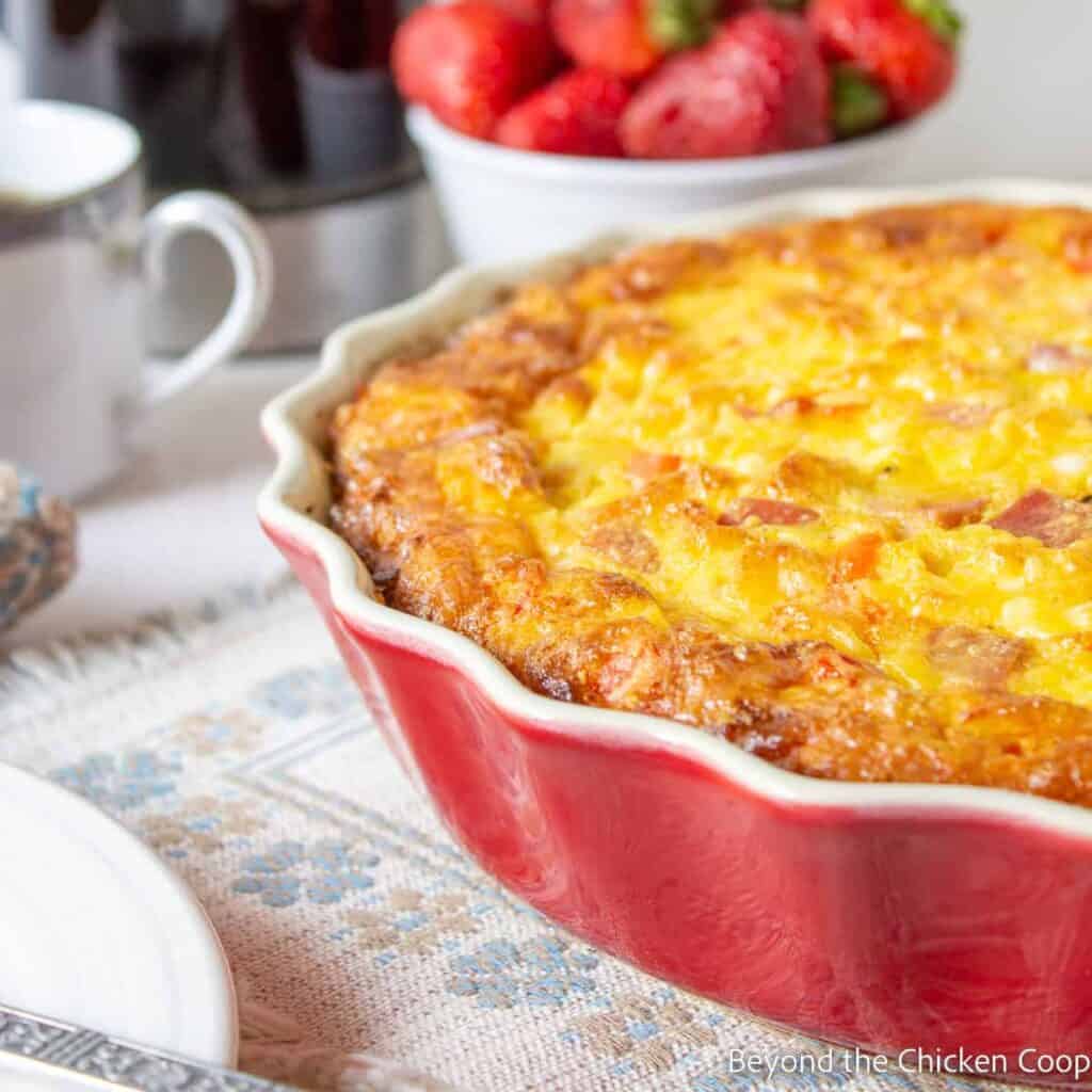 Egg casserole in a red pie dish on a staged breakfast table.