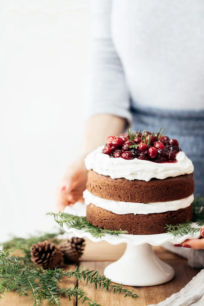 Two layered Gluten Free Gingerbread Cake on a cake plater with cranberries on top being placed on a staged wooden tabletop.
