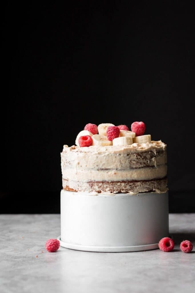 Double layered banana cake on a solid white cake stand topped with fresh raspberries and banana slices.