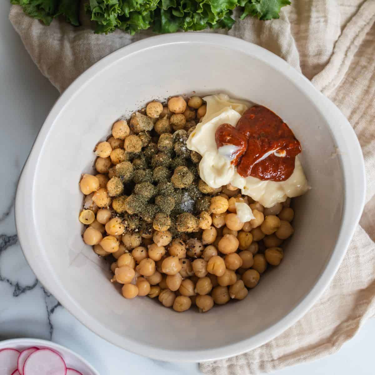 Chickpeas in a mixing bowl with spices, mayo and harissa on top.