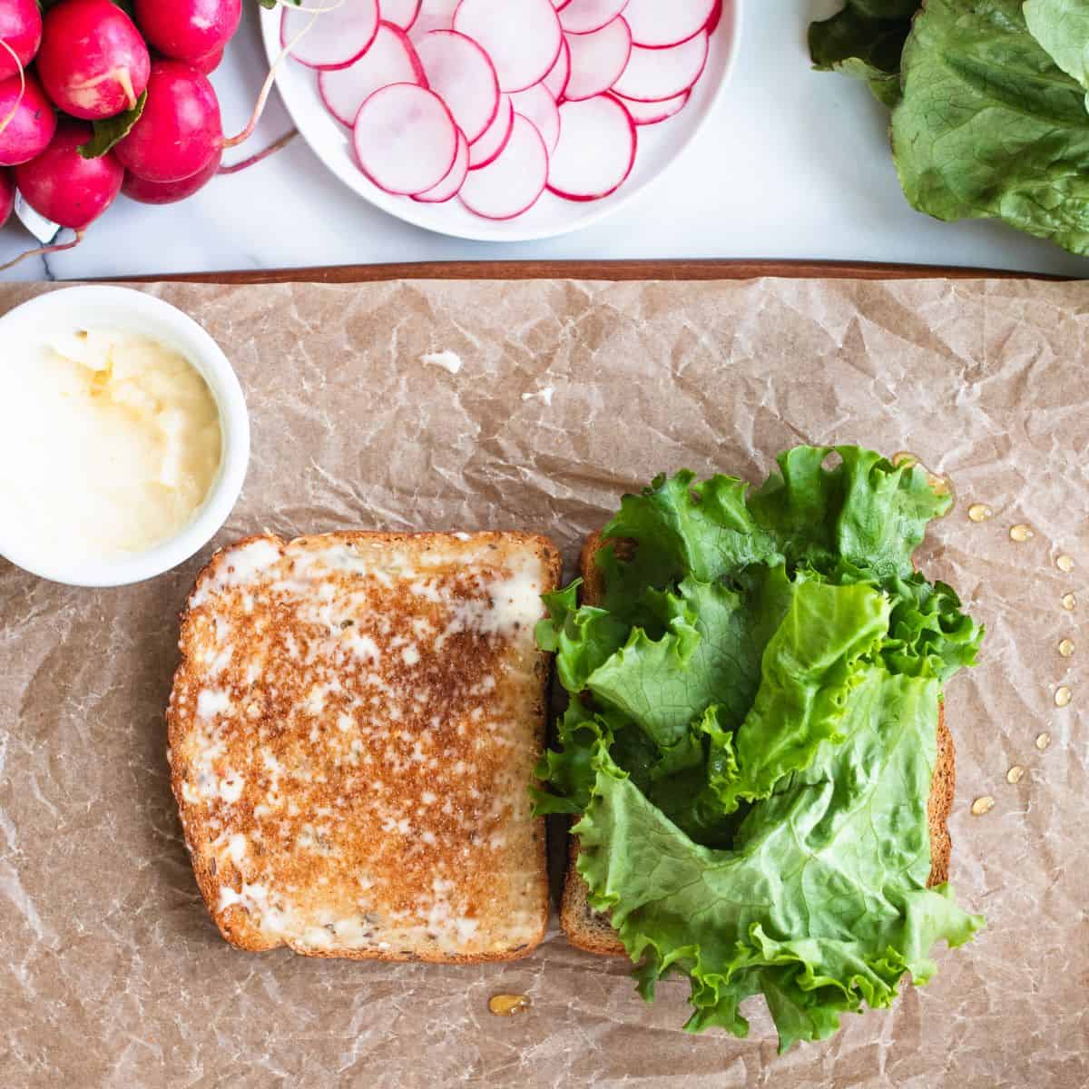 Toasted bread on parchment paper on a staged tabletop buttered, with big lettuce leaves on top.