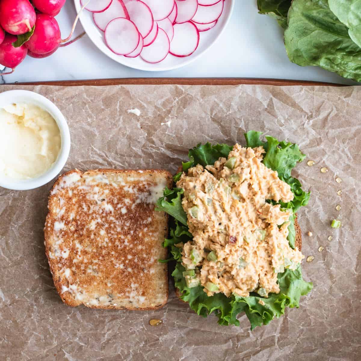 Toasted bread on parchment paper on a staged tabletop being buttered, with large lettuce leaves and chickpea smash on top.