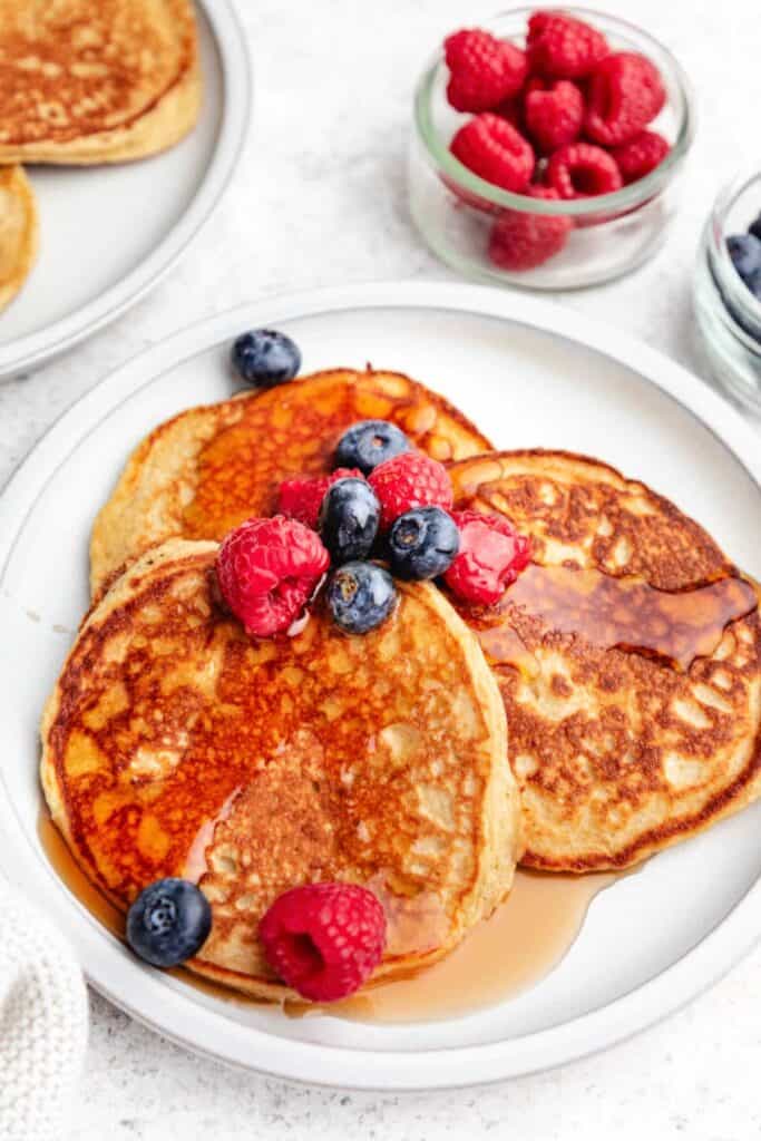 Three pancakes on a plate with fresh raspberries and blueberries and maple syrup on top with more fruit in small bowls beside.