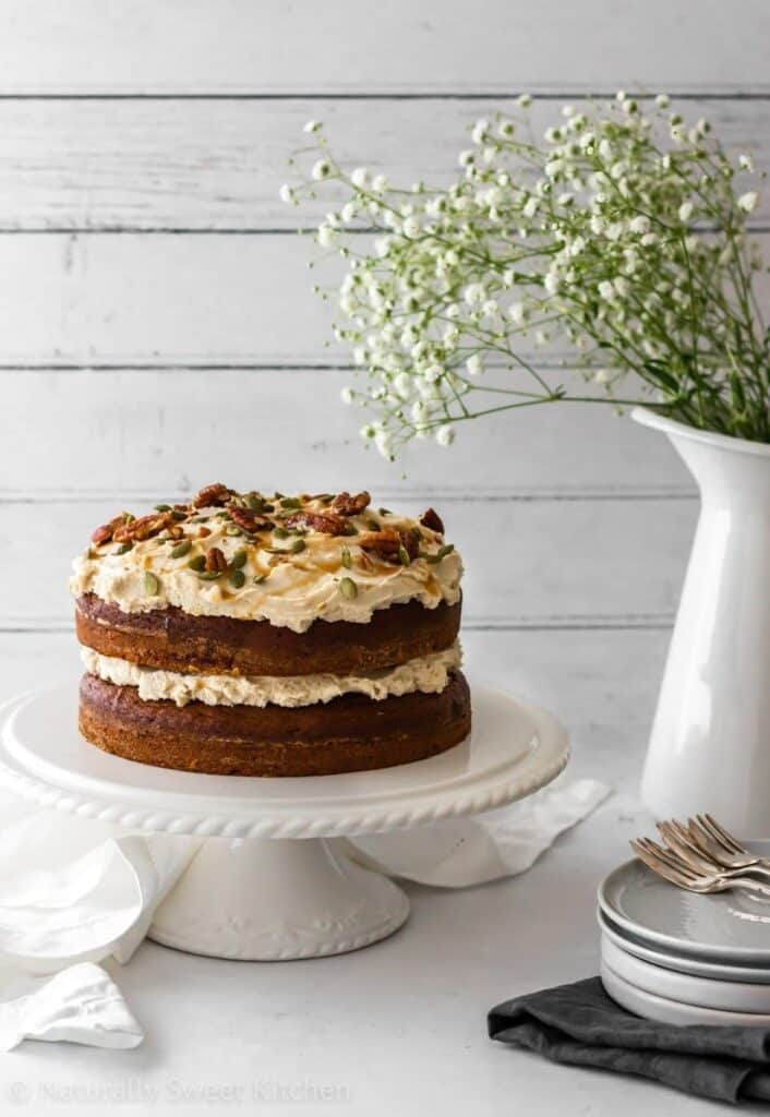 A double layered pumpkin spice cake on a cake stand with walnuts, pumpkin seeds and icing on top.