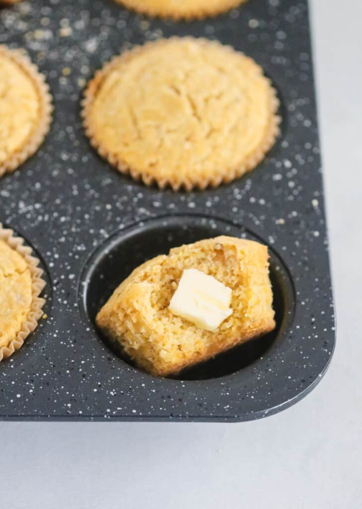 speckled muffin tin full of corn muffins with a focus on one with a bite out of it with a slice of butter in the bite.