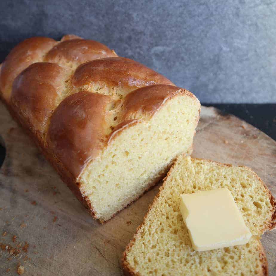 A slice of einkorn brioche on a table top with a slice of butter on it beside the loaf, all on a wooden cutting board.