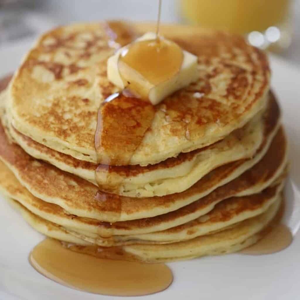 6 Einkorn pancakes stacked on top of each other with a cube of butter on top with maple syrup being poured on top.