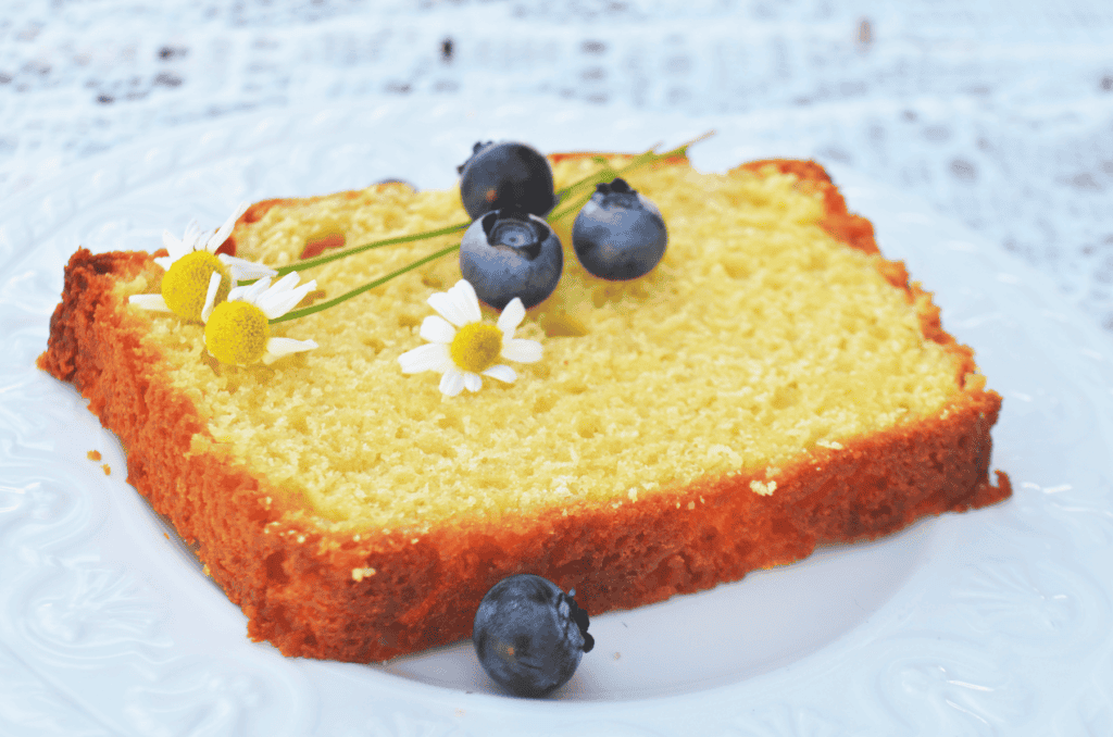 Piece of pound cake on a white plate with three daisies and blueberries on top.