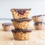 Three puffed quinoa bites stacked on each other on a tabletop full of them and the one on the has a bite out of it showing the jam center.