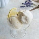 A dessert dish with two scoops of cottage cheese lemon ice cream and wedges of lemon on a staged tabletop with lavender on it.