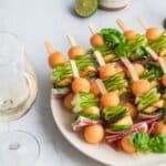 Halloumi skewers featuring melon balls on a serving plate beside a glass of white wine.